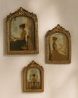Three framed vintage-style oil paintings depicting women at a window, arranged on a textured off-white wall under warm sunlight.