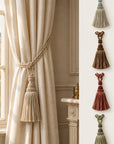 A cream-colored curtain tieback with a knotted rope cord and a large tassel is shown hanging on a window in an elegant room. On the right side, a vertical panel displays five different tassel tieback color options against a light wall.