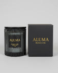 A black glass jar candle with a black lid and a black label reading ALUMA Blackberry & Bay, shown next to its matching black box with gold lettering on a light gray background.