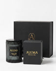 A black glass jar candle with a black lid and gold lettering, displayed alongside its black gift box and a smaller black presentation box with gold text. The brand name 'ALUMA' is clearly visible on the labels.