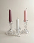 Three taper candles in clear glass candlesticks on a warm beige background: one dusty mauve, one white, and one soft pink.