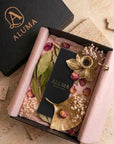An open black gift box lined with blush pink tissue paper, styled with dried pink roses, green leaves, and gold-toned decorative leaves. A black card and a black box lid with the gold ALUMA logo are placed inside and beside the box on a beige stone surface.