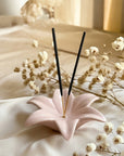 Pink ceramic incense holder with black sticks on a light fabric background with white flowers.