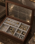 An open dark wood jewelry organizer box with a textured glass lid, filled with gold jewelry and arranged in compartments, set on a dark marble surface with books and a candle in the background.