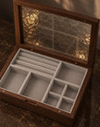 An open wooden jewelry organizer box with a textured glass lid, featuring light gray velvet interior compartments and ring rolls, set on a dark marble surface.