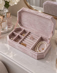 A blush pink velvet jewelry box with multiple compartments, shown open on a white marble vanity table with jewelry arranged inside and a mirror in the background.