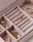 An open blush pink velvet jewelry organizer tray with multiple compartments, holding rings, earrings, and a pearl necklace.