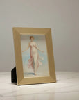 A luxury gold framed vintage-style painting of a woman in a flowing pale pink dress, displayed on a white marble surface against a light gray wall.