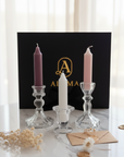 Three taper candles in glass candlesticks arranged on a marble surface in front of a black branded box. The candles are plum purple, white, and pale pink, with dried flowers and envelopes placed around them.