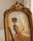 An ornate vintage-style gold picture frame holding a painted portrait of a woman in a yellow dress seated by a window, shown against a softly lit neutral interior background.