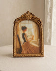 An ornate gold-framed vintage-style picture frame displaying a painting of a woman in a yellow dress by a window, set on a round table against a neutral cream wall and lace curtain background.