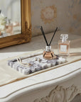 A styled interior scene showing a marble tray holding a reed diffuser and a small perfume bottle on an ornate cream console table, with a gold-framed mirror and floral wallpaper in the background.