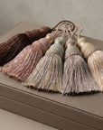 Five decorative tassels in earthy neutral tones including brown, dusty rose, beige, and cream, arranged on top of a textured taupe leather box.