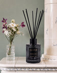 A reed diffuser bottle with black reeds sits on a white tiled console table next to a small vase of flowers, with a tall cream-colored ceramic column in the background.