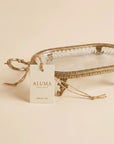 An oval clear glass tray with a decorative gold-toned metal rim and handles, shown with a hanging paper tag that reads ALUMA. The tray is photographed on a plain warm cream background.