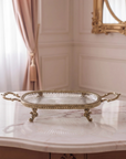 An ornate oval gold tray with a clear glass top and decorative handles, styled on a marble-topped console table with pearl necklaces draped across it in a classic interior setting.