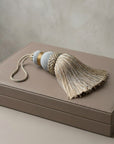 A decorative tassel with a cream fringe, a braided beige-and-white center, a light blue cylindrical section, and a small gold accent, resting on a taupe leather box against a neutral beige background.