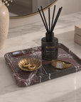 A reed diffuser bottle labeled ALUMA with black reeds, displayed on a dark marble tray alongside gold jewelry and a watch on a light marble countertop.
