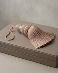 A dusty pink and beige embroidered tassel with a matching cord lies on top of a taupe leather jewelry box against a neutral textured background.
