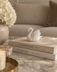 ceramic white swan on top of tow home decor books, on top of a white marble table, next to flower vase, cream background and cream sofa at the conner.