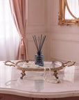 A reed diffuser bottle with black reeds sits on an ornate gold tray draped with a pearl necklace, placed on a marble table in a softly lit room with pink curtains and a gold mirror.