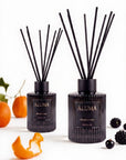 Two ALUMA reed diffusers with black glass bottles and black reeds, styled with citrus peels, oranges, and blackberries on a white background.