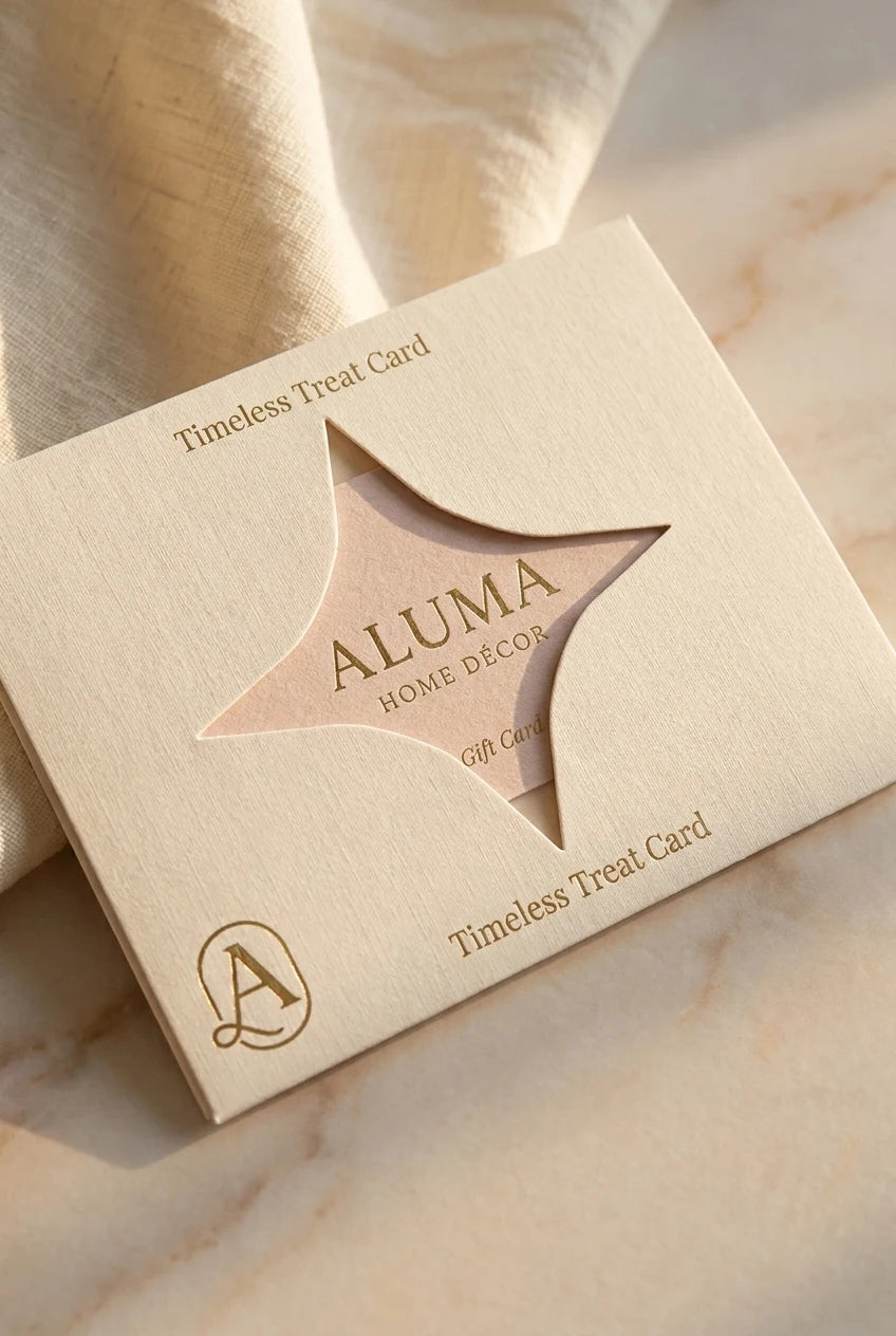 A square cream-colored gift card box from ALUMA Home Decor, featuring a star-shaped cutout that reveals a pinkish-beige card inside. The box is printed with 'Timeless Treat Card' and a monogram logo, resting on a soft beige fabric and marble surface.