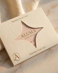 A square cream-colored gift card box from ALUMA Home Decor, featuring a star-shaped cutout that reveals a pinkish-beige card inside. The box is printed with 'Timeless Treat Card' and a monogram logo, resting on a soft beige fabric and marble surface.