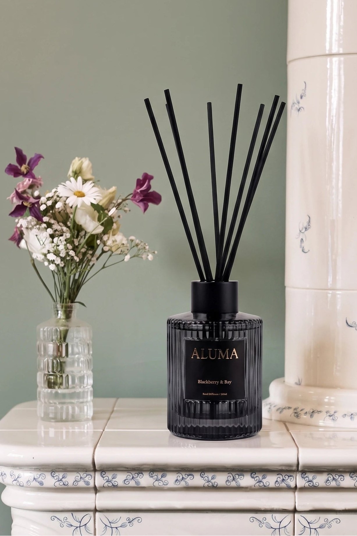 A reed diffuser bottle with black reeds sits on a white tiled console table against a sage green wall, styled with a small glass vase of flowers and a large cream ceramic column.
