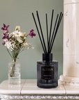 A reed diffuser bottle with black reeds sits on a white tiled console table against a sage green wall, styled with a small glass vase of flowers and a large cream ceramic column.