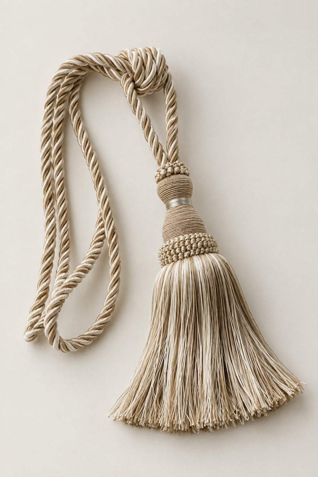 A decorative beige and cream tassel with a twisted rope cord, knotted top, and textured beaded band, photographed on a plain light beige background.