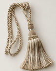 A decorative beige and cream tassel with a twisted rope cord, knotted top, and textured beaded band, photographed on a plain light beige background.