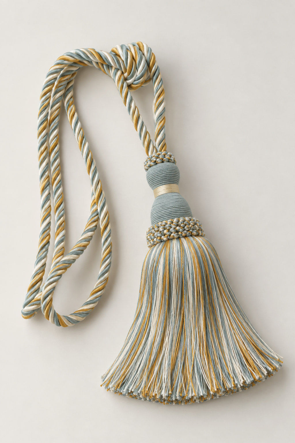 A decorative tassel with a braided cord and a fringed tassel, featuring a muted blue-gray and mustard yellow striped pattern, accented with a beaded band and a gold-toned collar, set against a plain light beige background.