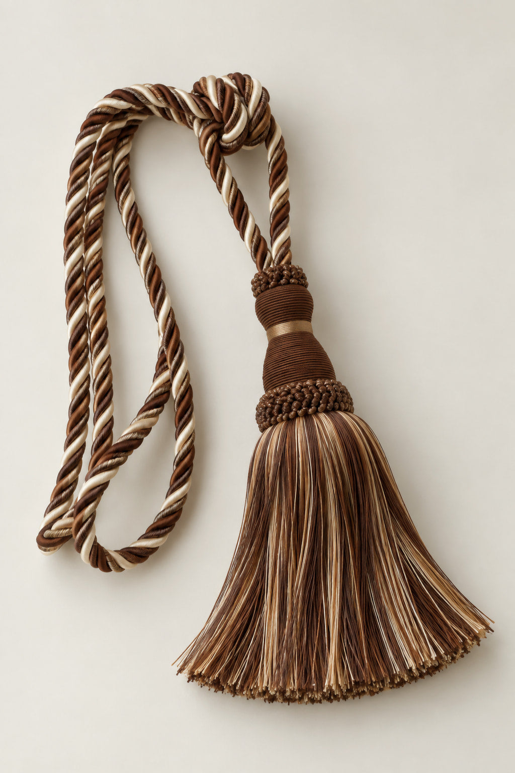 A decorative curtain tassel with twisted braided cords in brown, beige, and cream tones, photographed on a plain light background.