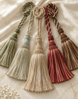 Four decorative curtain tassels with braided cords and long fringe, arranged on a soft cream fabric background. The tassels come in muted sage green, pale blue, cream, and burgundy-red tones.