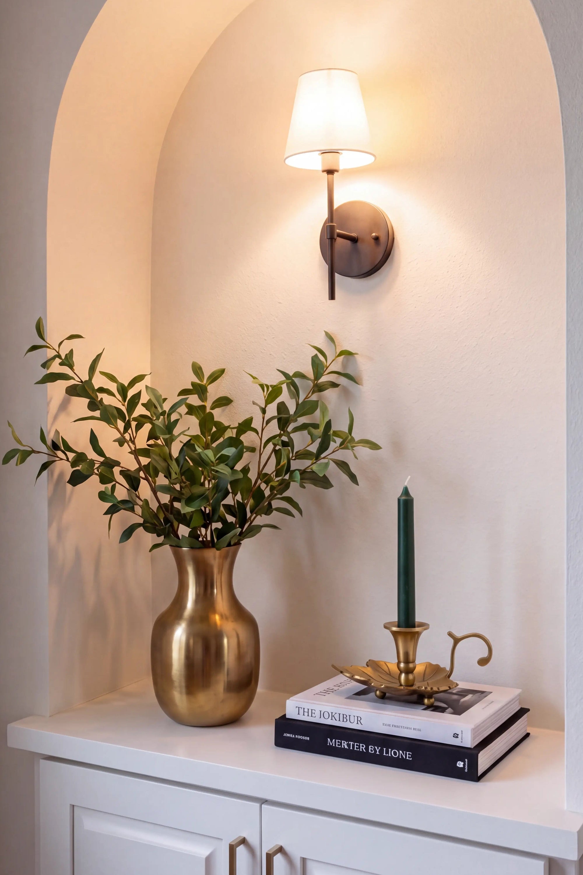 A warm wall sconce with a white drum shade and dark metal mount is mounted on an arched beige wall above a white console table. The table is styled with a gold vase of green leaves, a stack of books, and a brass candlestick with a green taper candle.