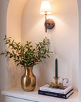 A warm wall sconce with a white drum shade and dark metal mount is mounted on an arched beige wall above a white console table. The table is styled with a gold vase of green leaves, a stack of books, and a brass candlestick with a green taper candle.