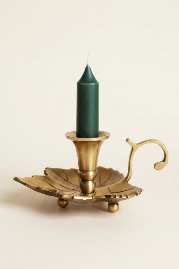 A brass leaf-shaped candle holder with a curved handle, holding a dark green taper candle, set against a plain off-white background.