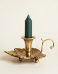 A brass leaf-shaped candle holder with a curved handle, holding a dark green taper candle, set against a plain off-white background.