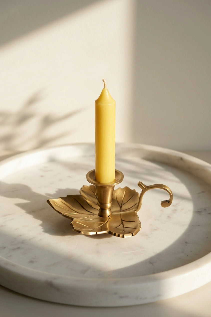 A yellow taper candle in a brass gold leaf-shaped metal holder, styled on a round white marble tray with warm natural sunlight and leaf shadows.