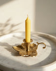 A yellow taper candle in a brass gold leaf-shaped metal holder, styled on a round white marble tray with warm natural sunlight and leaf shadows.
