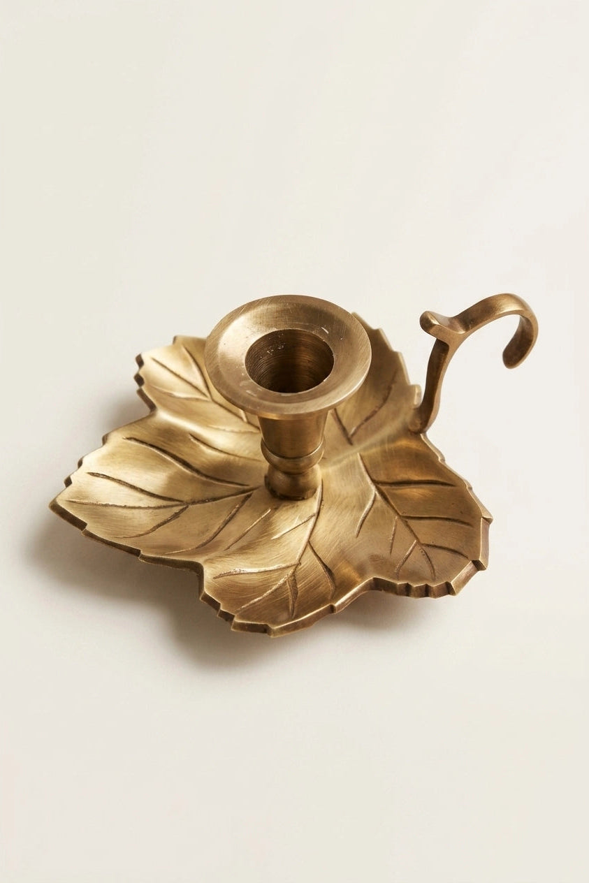 A brass leaf-shaped candle holder with a central cup and a curved hook handle, shown on a plain light background.