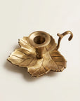 A brass leaf-shaped candle holder with a central cup and a curved hook handle, shown on a plain light background.