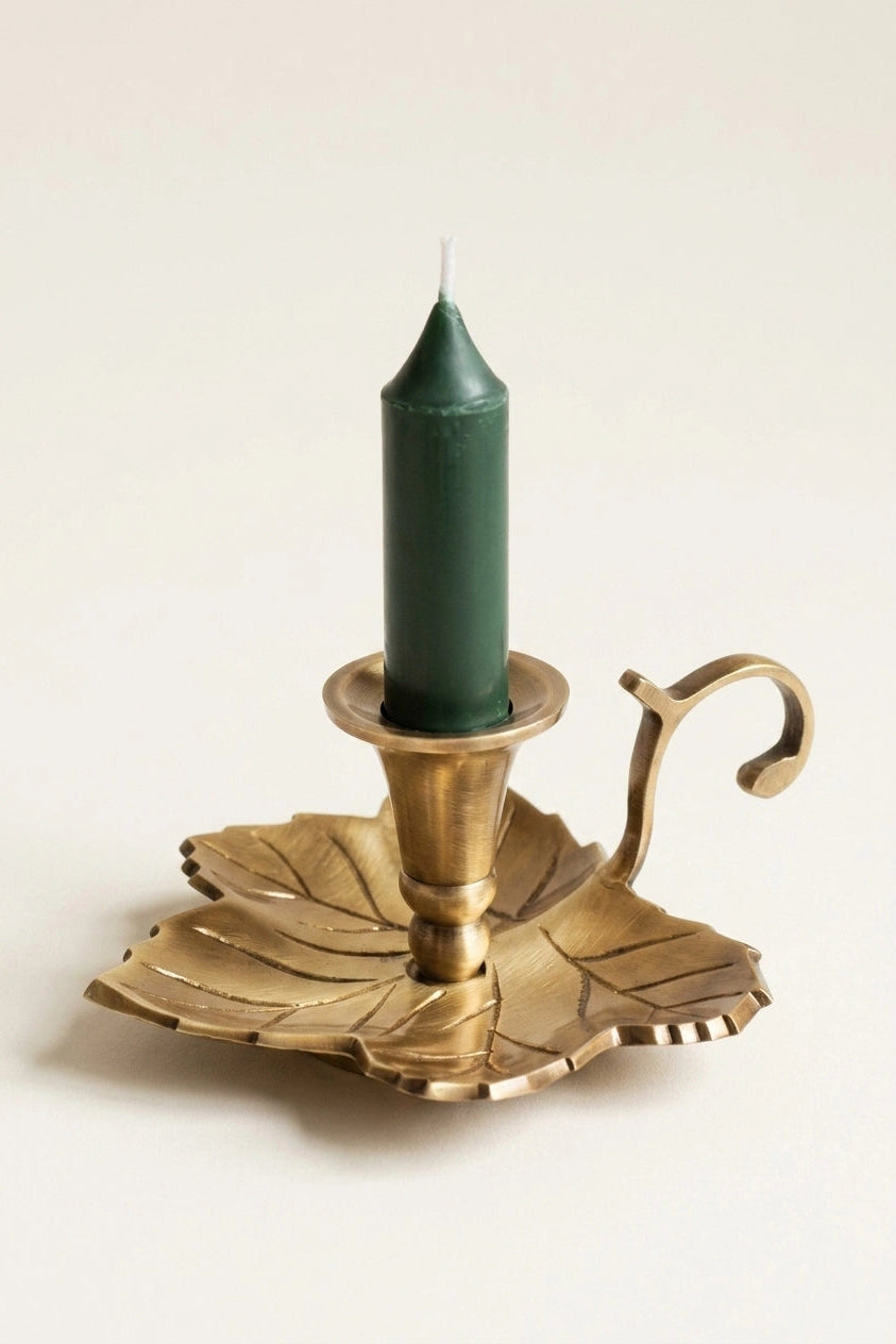 A brass leaf-shaped candle holder with a curved handle, holding a dark green taper candle, set against a plain off-white background.