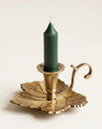 A brass leaf-shaped candle holder with a curved handle, holding a dark green taper candle, set against a plain off-white background.