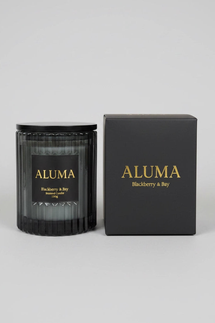 A black glass jar candle with a black lid and a black label reading ALUMA Blackberry & Bay, shown next to its matching black box with gold lettering on a light gray background.