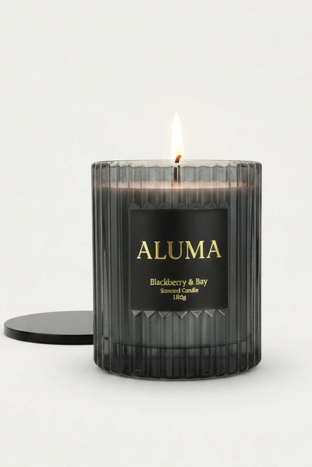 A lit ALUMA candle in a ribbed dark glass jar with a black label and gold lettering, shown on a clean white background with its black lid beside it.