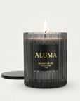 A lit ALUMA candle in a ribbed dark glass jar with a black label and gold lettering, shown on a clean white background with its black lid beside it.