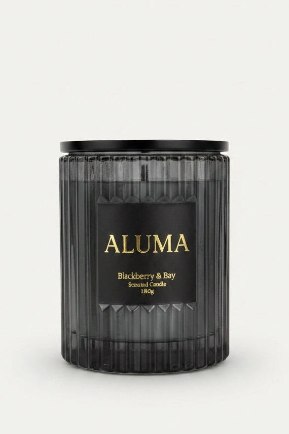 A dark charcoal gray ribbed glass candle jar with a black lid and a black label featuring gold lettering that reads 'ALUMA' and 'Blackberry & Bay'.