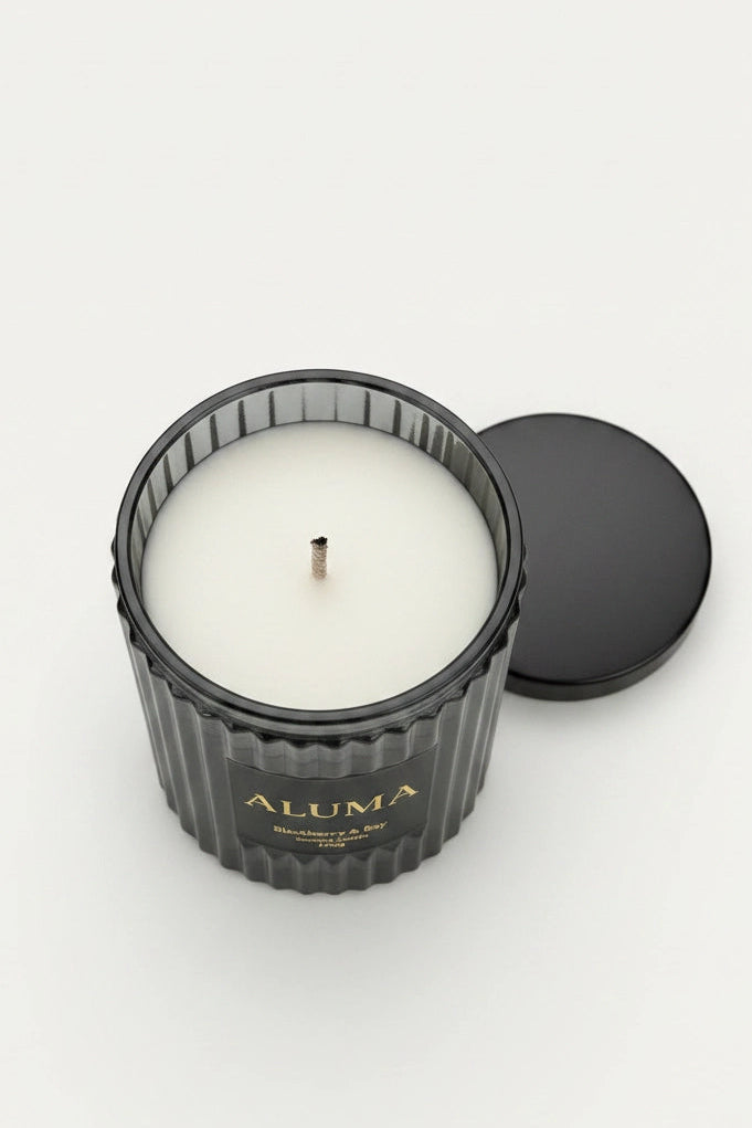An open black ribbed glass candle jar with white wax and a single wick, shown on a light gray background with its black lid beside it. The jar is labeled ALUMA.